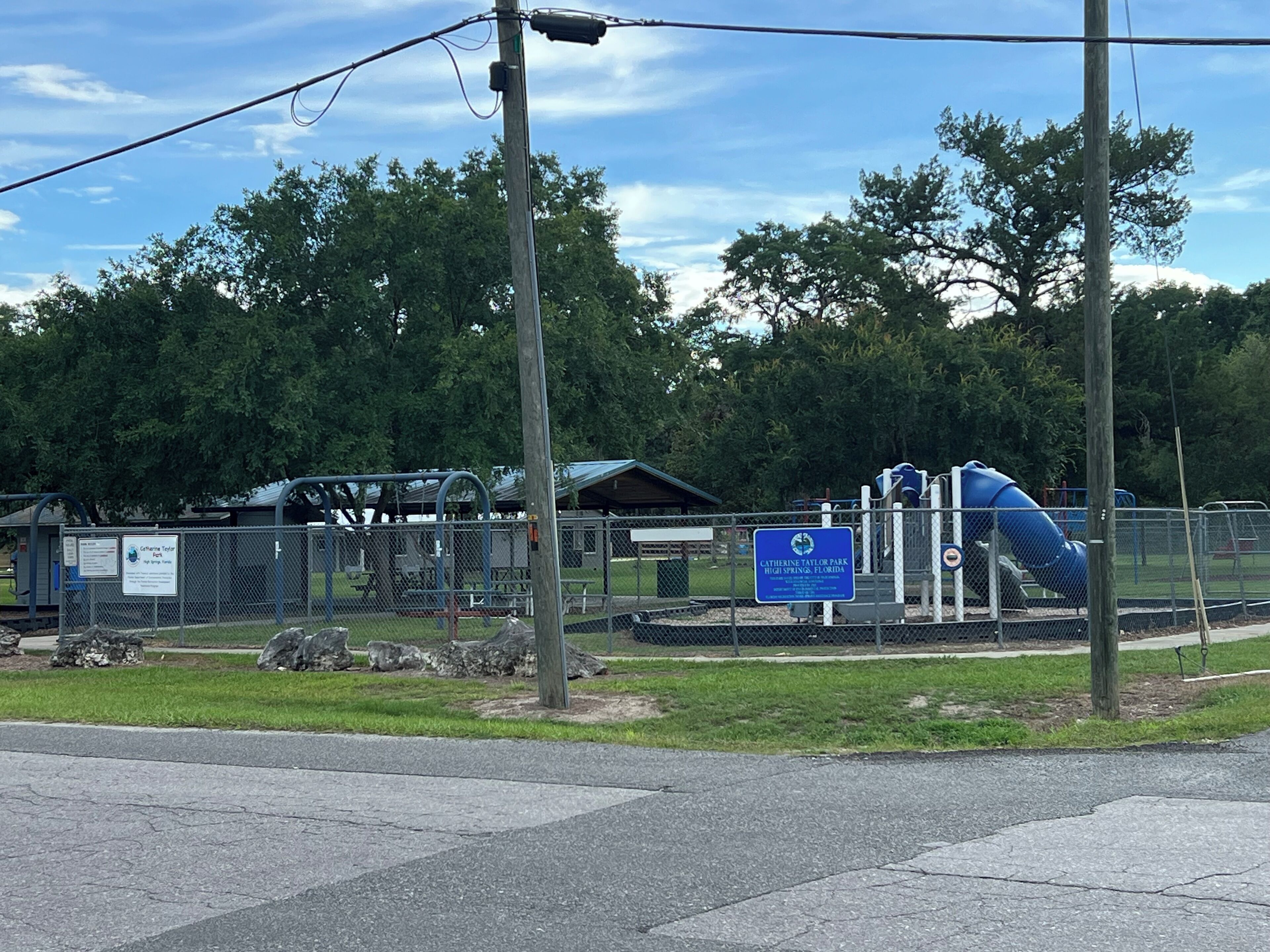 A fenced playground area with a blue slide and climbing structures, surrounded by trees and a picnic shelter.