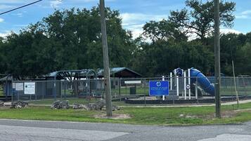A fenced playground area with a blue slide and climbing structures, surrounded by trees and a picnic shelter.