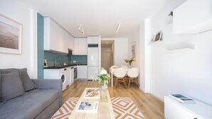 A modern living space with a grey sofa, a dining area with a table and chairs, a kitchen with white cabinets, and a small plant on a shelf. - Edmond Vibes Airport - Ifema Hub (Madrid)