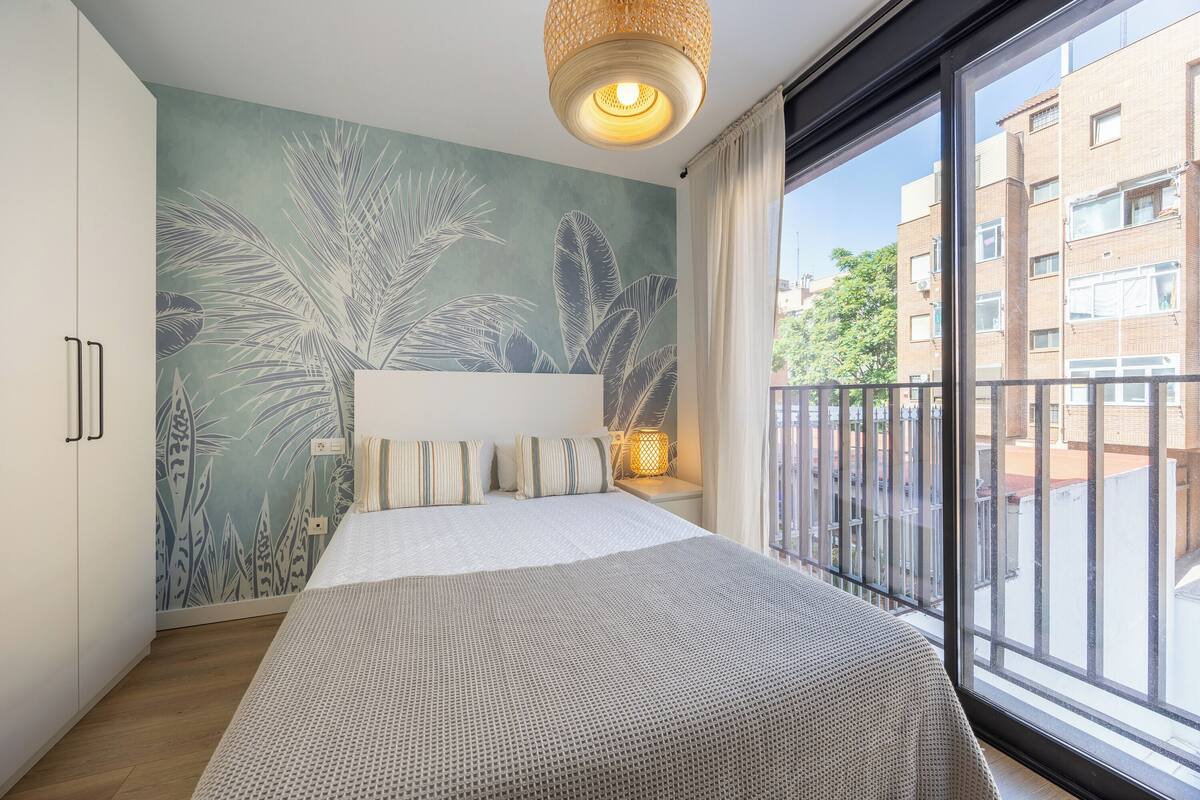 Un dormitorio con cama, un mural en la pared, una ventana con vista a edificios y un balcón.