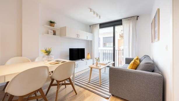 A modern living room with a grey sofa, a dining table with chairs, a TV, and a small table with a vase and book. - Edmond Vibes Airport - Ifema Hub (Madrid)