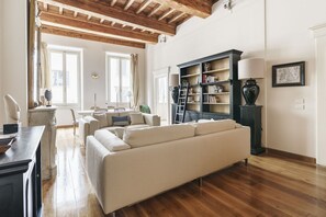 A spacious living room with a large beige sofa, wooden flooring, a bookshelf, and a lamp.