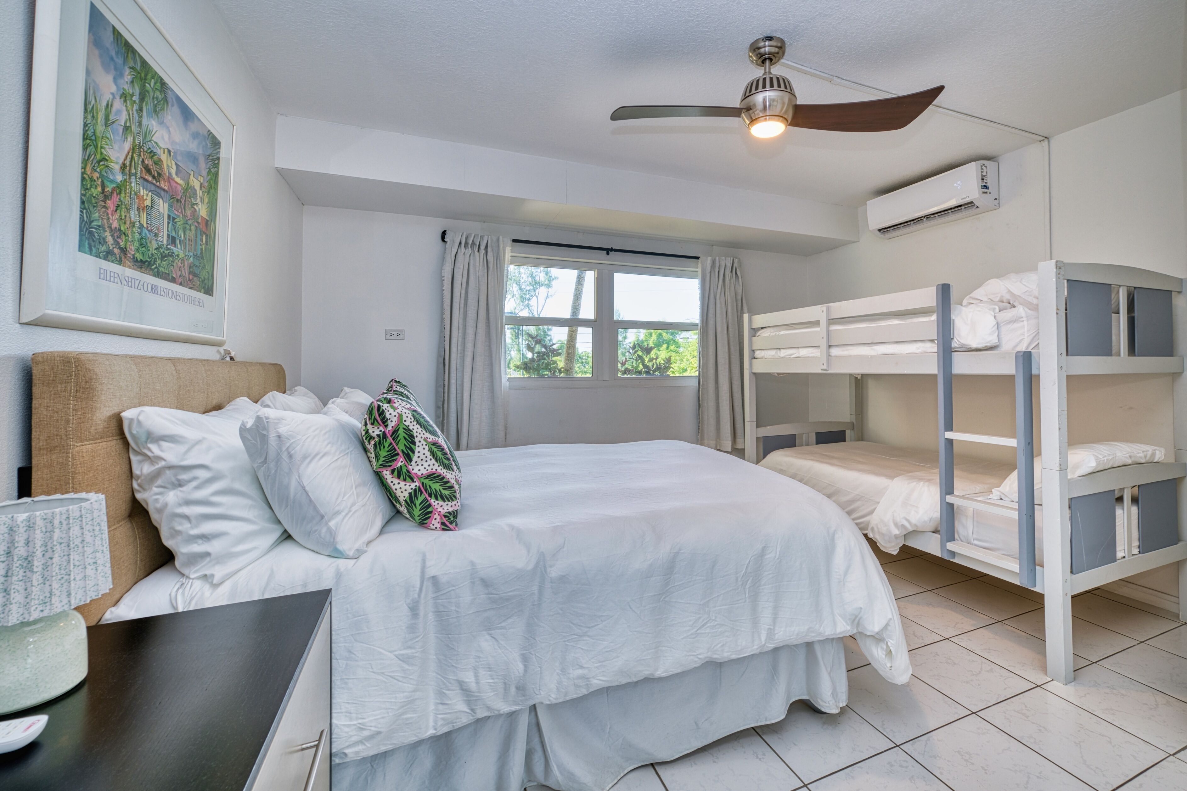 Ein Zimmer mit einem Etagenbett, einem Einzelbett, einem Deckenventilator, einer Klimaanlage, einem Fenster mit Vorhängen und einem gerahmten Bild an der Wand.