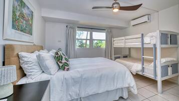 Ein Zimmer mit einem Etagenbett, einem Einzelbett, einem Deckenventilator, einer Klimaanlage, einem Fenster mit Vorhängen und einem gerahmten Bild an der Wand.