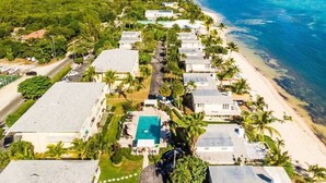 An aerial view of a coastal resort with multiple bungalow-style buildings, a central swimming pool, and a beach adjacent to the ocean. - Quiet Beach Escape - Caribbean Paradise (South Sound)