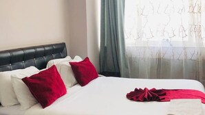 A hotel room with a bed, white bedding, red pillows, a black headboard, and a window with sheer curtains. - Shalla Guesthouse and Wellness Center (Addis Ababa)