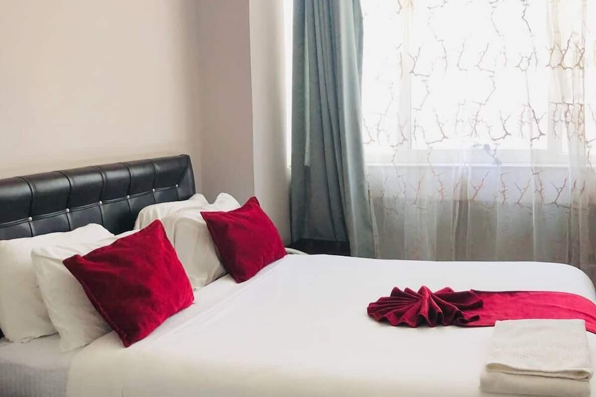 A hotel room with a bed, white bedding, red pillows, a black headboard, and a window with sheer curtains.
