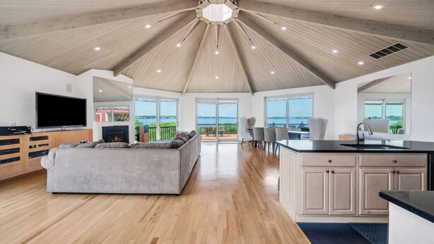 A spacious living room with a large gray sofa, a flat-screen TV, and a fireplace. The room features wooden flooring, a high ceiling with wooden beams, and large windows offering a view of the sea.