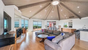 A spacious living area with a large wooden ceiling, a fireplace, a sofa, and a view of the sea through large windows.