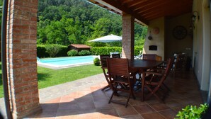 Un patio techado con un pilar de ladrillo, una mesa con sillas y vistas a una piscina y abundante vegetación.