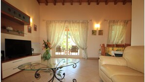Una sala de estar con una mesa de centro de vidrio, un sofá color beige, un televisor sobre un mueble de madera y vistas a una zona exterior con mobiliario a través de una puerta de vidrio.