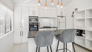 Une cuisine moderne avec des meubles blancs, un îlot central doté de deux tabourets de bar gris, un four encastré, un four à micro-ondes et un réfrigérateur.