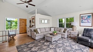 Un salon spacieux avec un grand canapé d’angle, une table basse et un tapis à motifs. On y trouve une porte en bois et une fenêtre donnant sur un paysage verdoyant.
