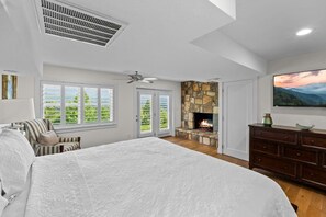 A spacious bedroom with a large bed, a stone fireplace, a ceiling fan, and a view of greenery through the windows.
