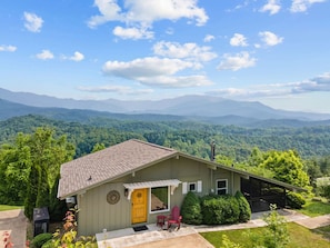 A small, single-story building with a sloped roof, a front porch, and a garage. It is surrounded by lush greenery and mountains in the background.