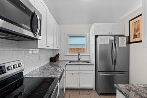 A modern kitchen with white cabinets, a stainless steel refrigerator, a built-in oven, and a window with blinds.
