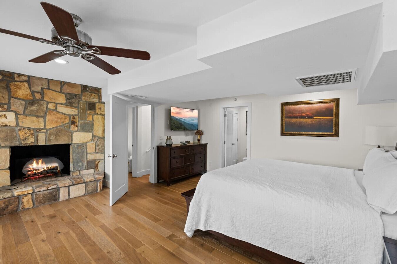 A bedroom with a stone fireplace, a bed, a ceiling fan, and a painting on the wall.