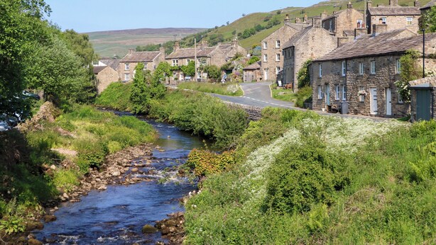 A stone-built village with a narrow river running through it, surrounded by greenery and hills in the background.