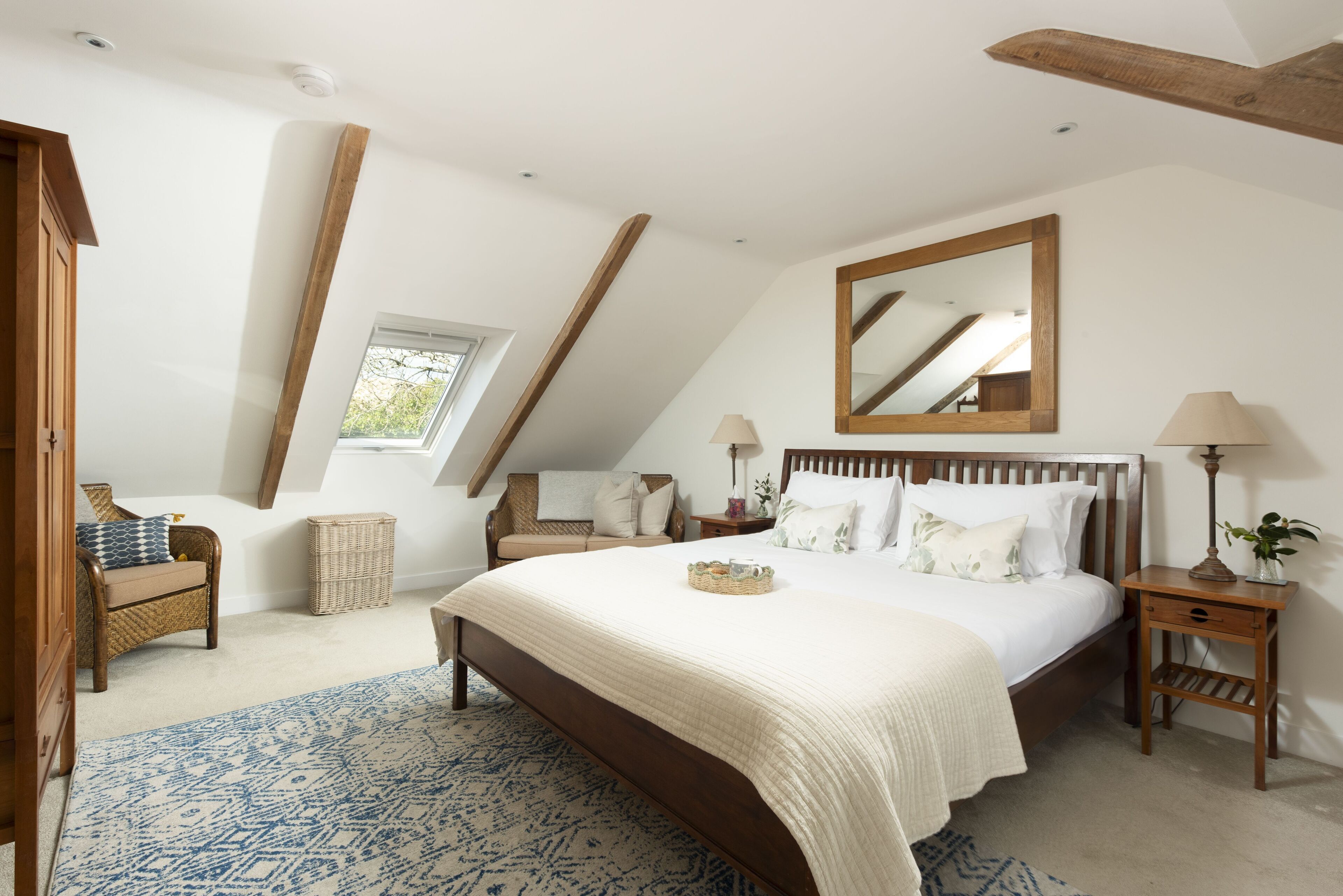 Ein Schlafzimmer mit einem großen Bett, einem Holzkopfstück, einem Nachttisch, einer Lampe, einem Korbstuhl, einem Fenster und einer schrägen Decke.