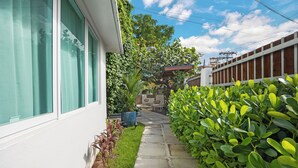 Un sendero conduce a una casa de fachada blanca, con un césped verde y diversas plantas.