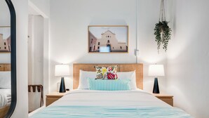 Habitación de hotel con cama, dos mesitas de noche, una planta colgante, un cuadro enmarcado en la pared y una ventana con vistas a edificios.