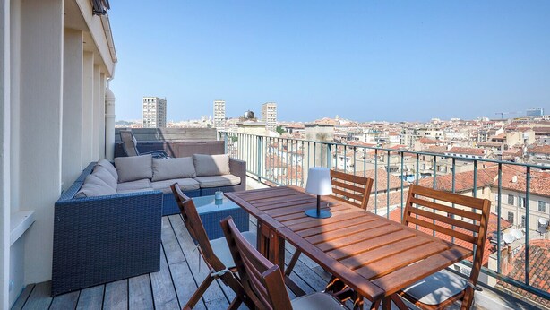 A balcony with a wicker sofa, wooden table, and chairs, overlooking a cityscape. - Cosy studio with terrace overlooking Marseille (Marseille)