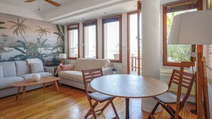 A living room with a sofa, a coffee table, a dining table with chairs, a ceiling fan, and large windows with wooden frames. - Cosy studio with terrace overlooking Marseille (Marseille)