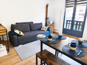 Ein modernes Wohnzimmer mit Sofa, Esstisch mit Unterlagen, einer Bank, einem kleinen Beistelltisch mit Lampe und Blick durch eine Tür.