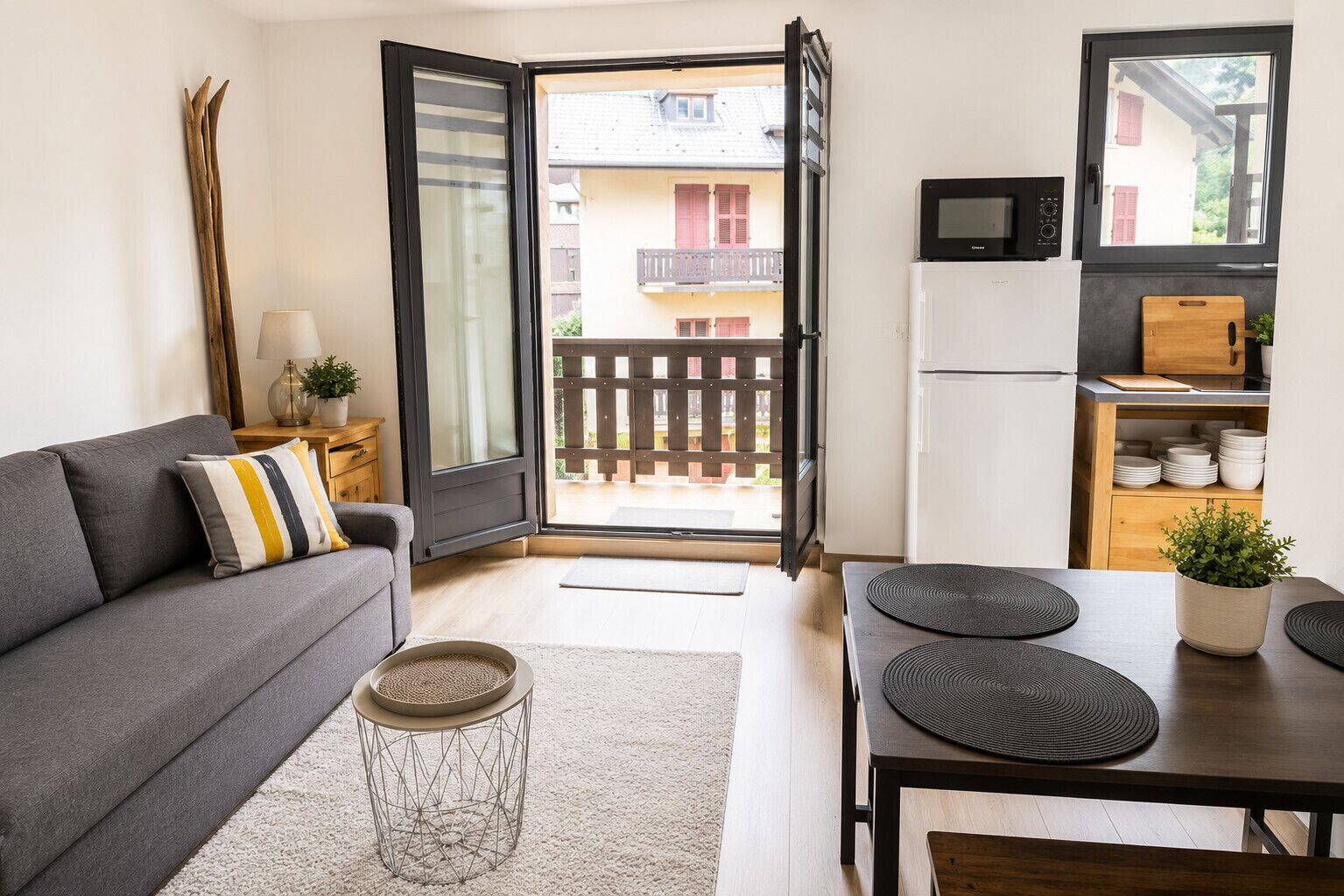 Een modern appartement met een grijze bank, een eettafel met zwarte placemats, een keuken met een magnetron en uitzicht op een balkon met een houten hek.
