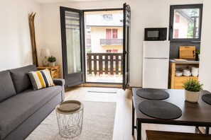 A modern apartment with a grey sofa, a dining table with black placemats, a kitchen with a microwave, and a view of a balcony with a wooden fence.