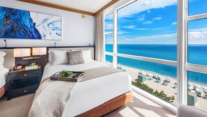 Une chambre avec une grande fenêtre donnant sur une plage, un lit avec du linge de lit blanc, une table de chevet avec une lampe et un tableau accroché au mur.