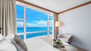 Une chambre d’hôtel dotée d’une grande fenêtre donnant sur l’océan, d’un lit avec du linge de lit blanc, d’une table de chevet avec un plateau et d’une lampe.