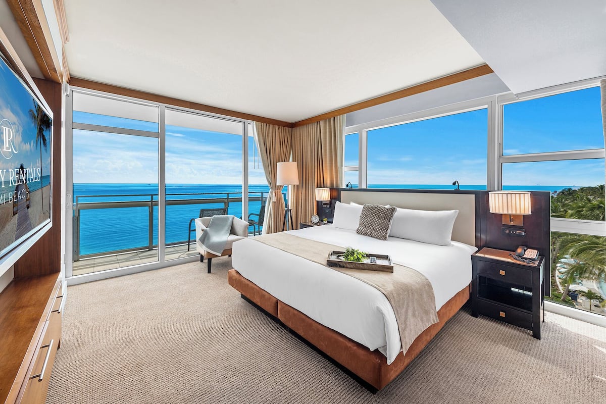 A spacious bedroom with a large bed, a nightstand, a chair, and a view of the ocean through floor-to-ceiling windows.