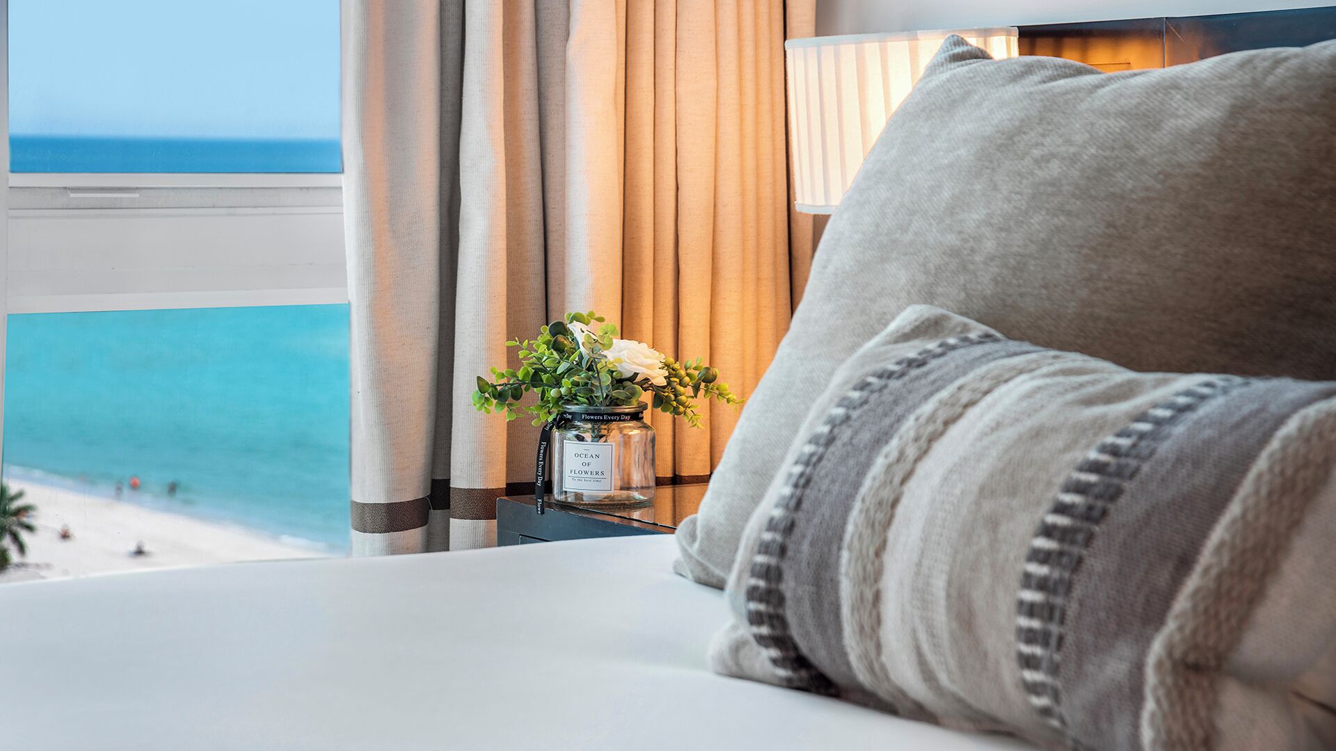 A hotel room with a view of the beach, a bedside table with a vase of flowers, and a lamp.