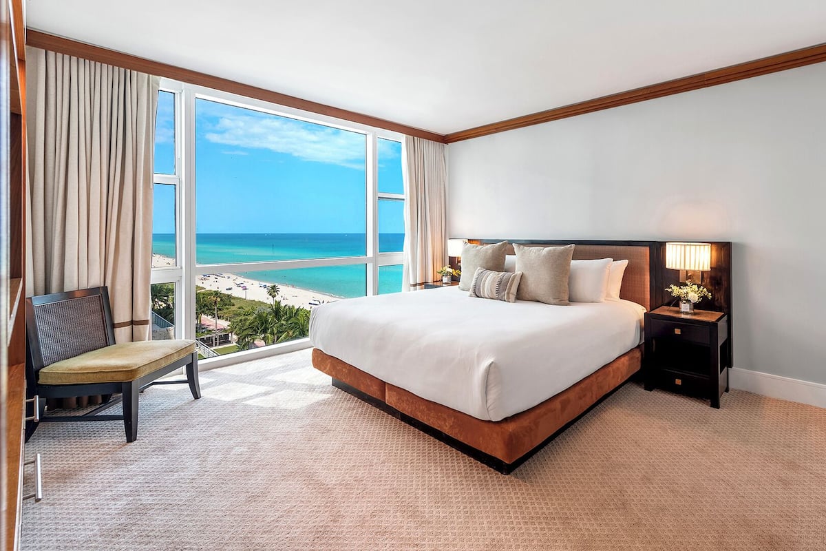 A hotel room with a large bed, a chair, a nightstand with a lamp, and a view of the beach and ocean.
