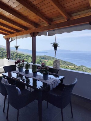 A covered outdoor dining area with a table and chairs, potted plants, and a view of the sea and hills.