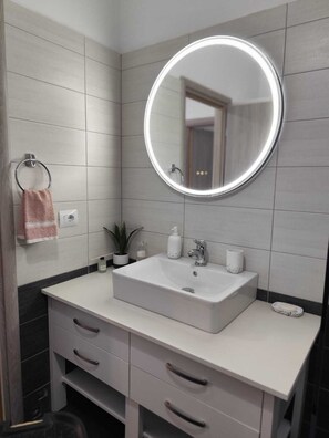 A modern bathroom with a white sink, a round mirror with a light, a towel ring, a potted plant, and a small cabinet.
