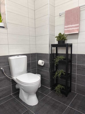 A modern bathroom with a white toilet, dark grey tiles, a black shelf with plants, and a pink towel on a rack.