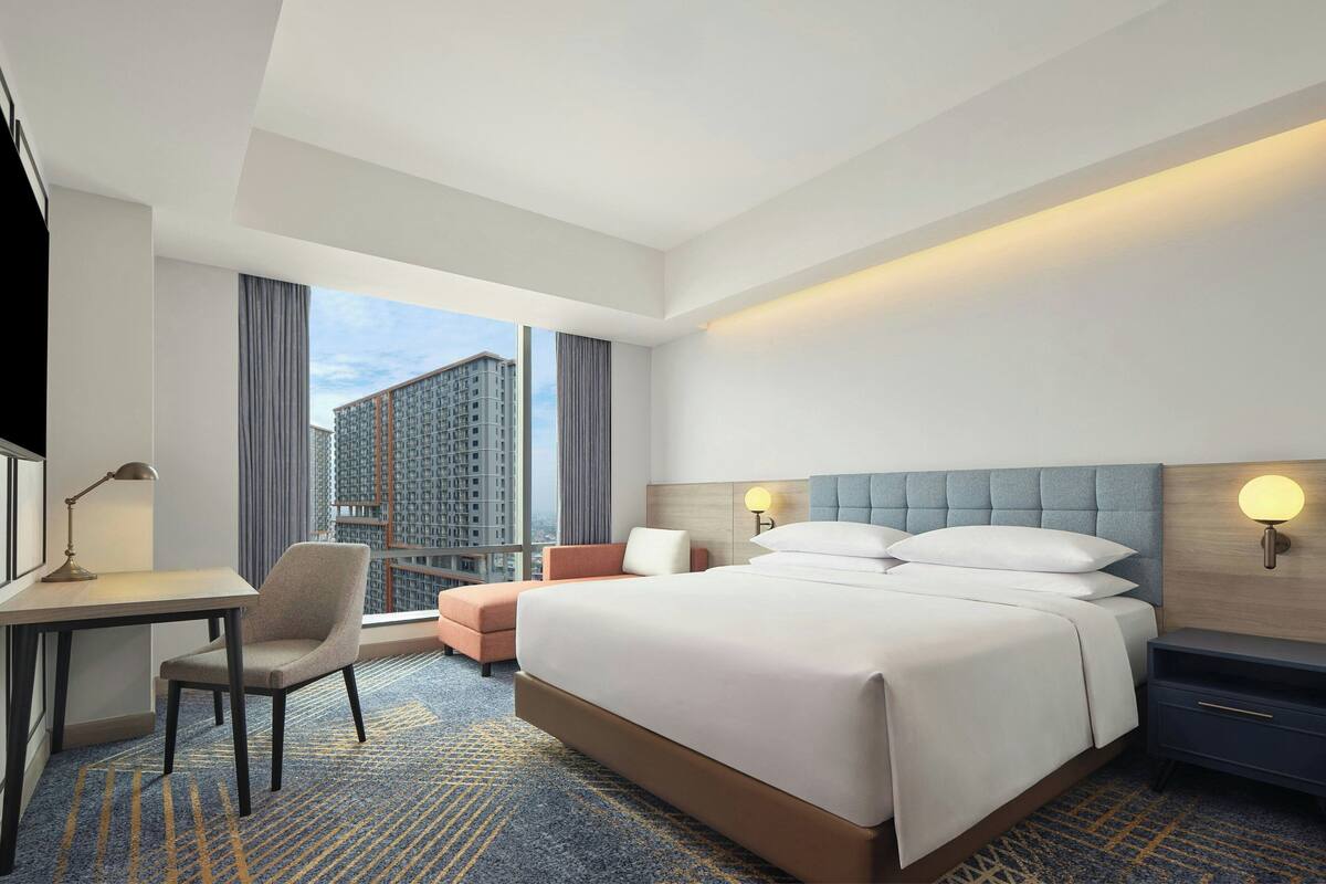 Une chambre d’hôtel avec un grand lit, un bureau, une chaise et une vue sur de hauts immeubles.