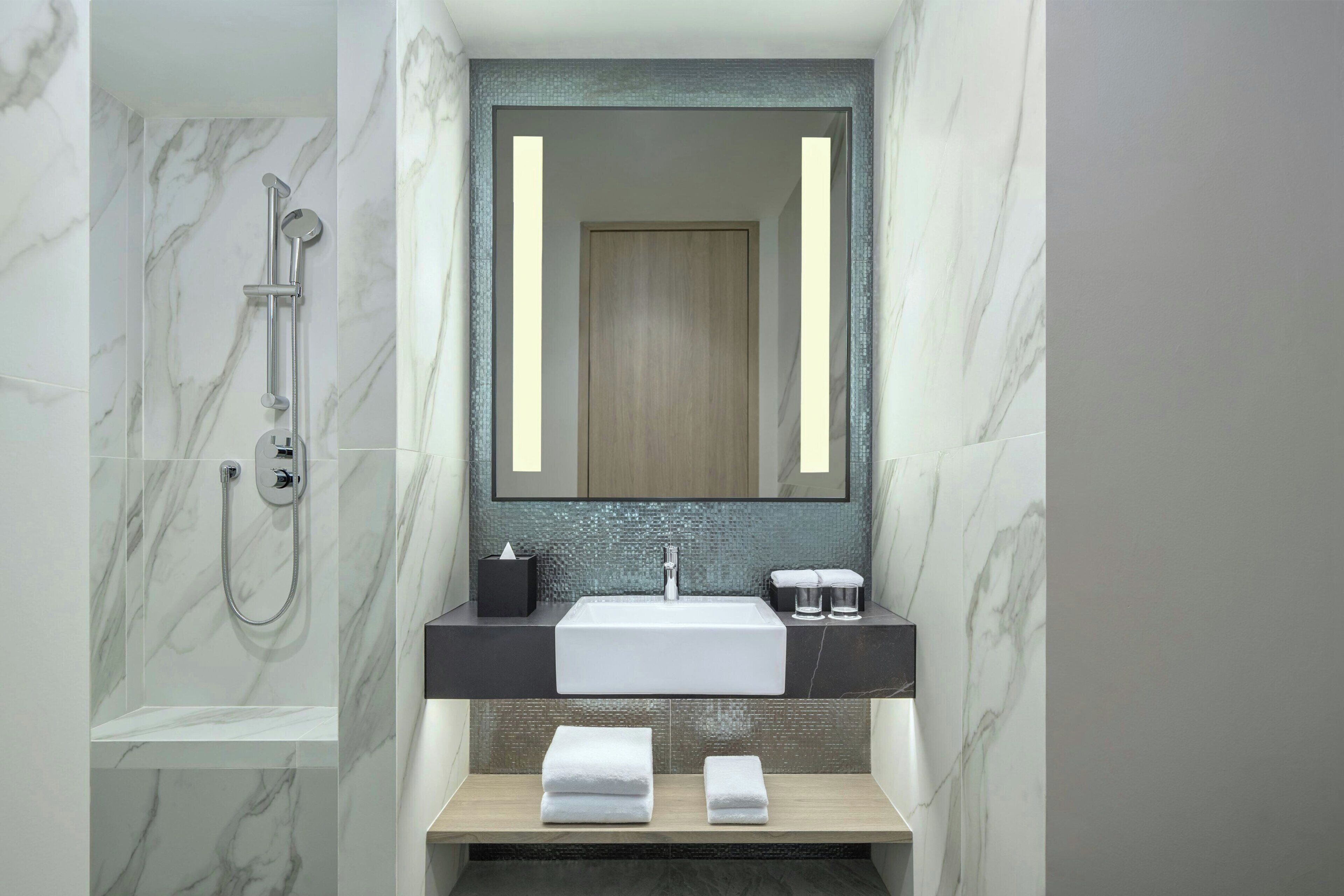 Une salle de bain moderne avec une douche en marbre, un grand miroir, un lavabo et deux serviettes posées sur une étagère.