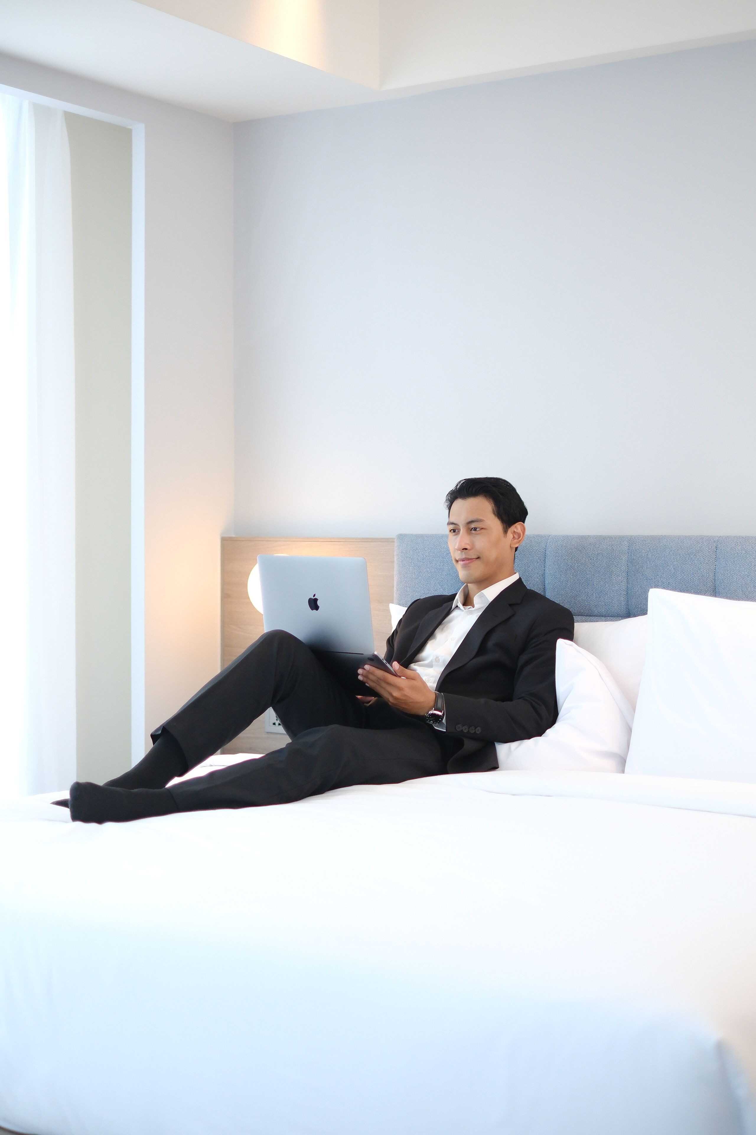 Een man in een zwart pak zit op een wit bed met een laptop, in een kamer met een lichtblauwe hoofdeinde en een bedlampje.