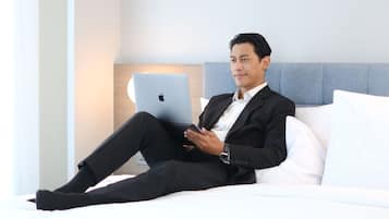 Een man in een zwart pak zit op een wit bed met een laptop, in een kamer met een lichtblauwe hoofdeinde en een bedlampje.