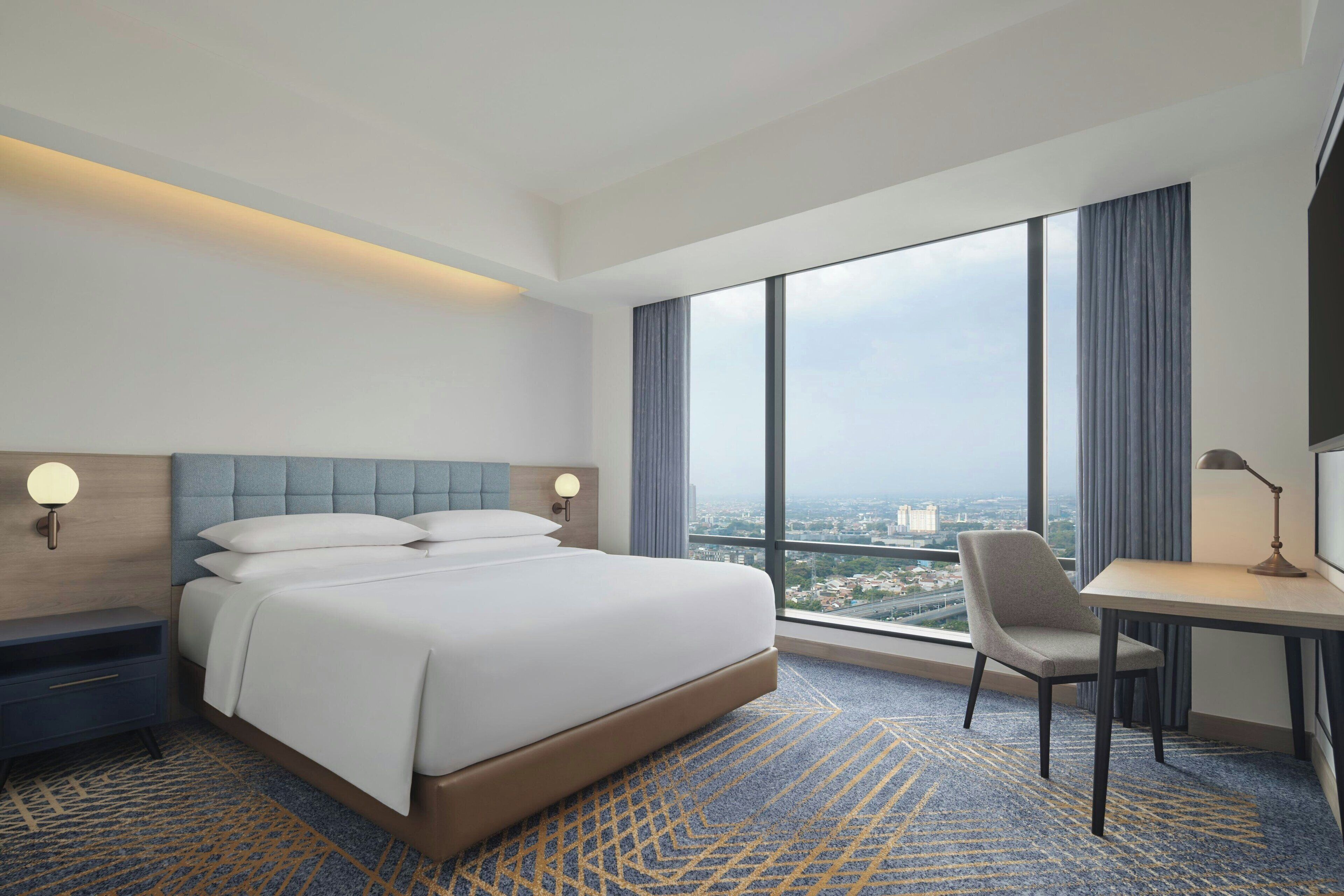 A hotel room with a large bed, a desk, and a chair. There is a view of the city through large windows.