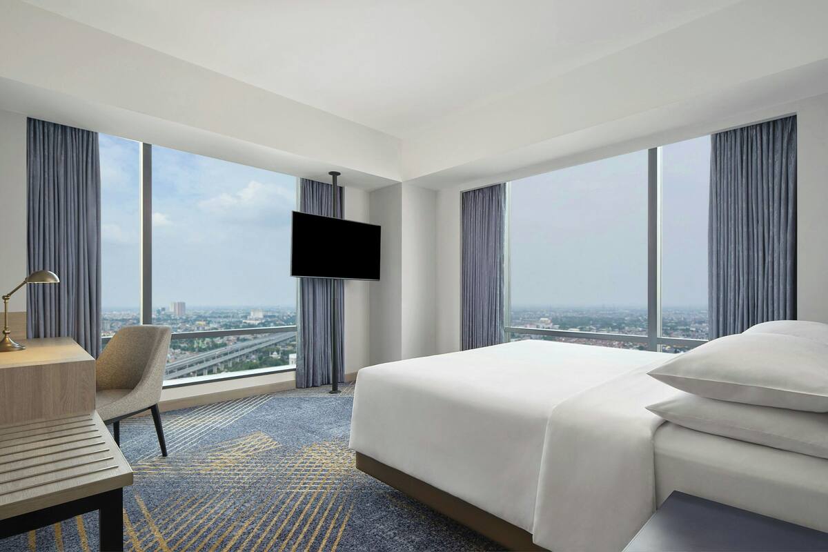 Une chambre d’hôtel avec un grand lit, un bureau, une télévision et une vue sur le paysage urbain grâce à de grandes fenêtres.