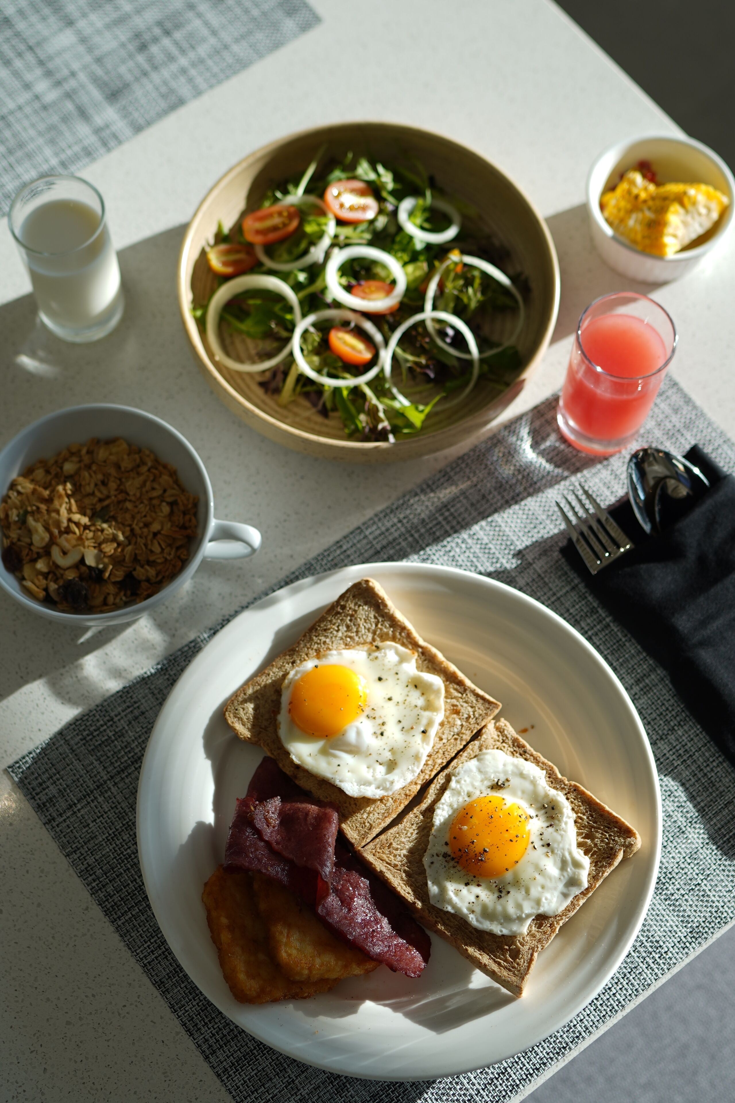 Una mesa de desayuno con tostadas, huevos, tocino, un tazón de ensalada, un vaso de jugo y un tazón de cereal.