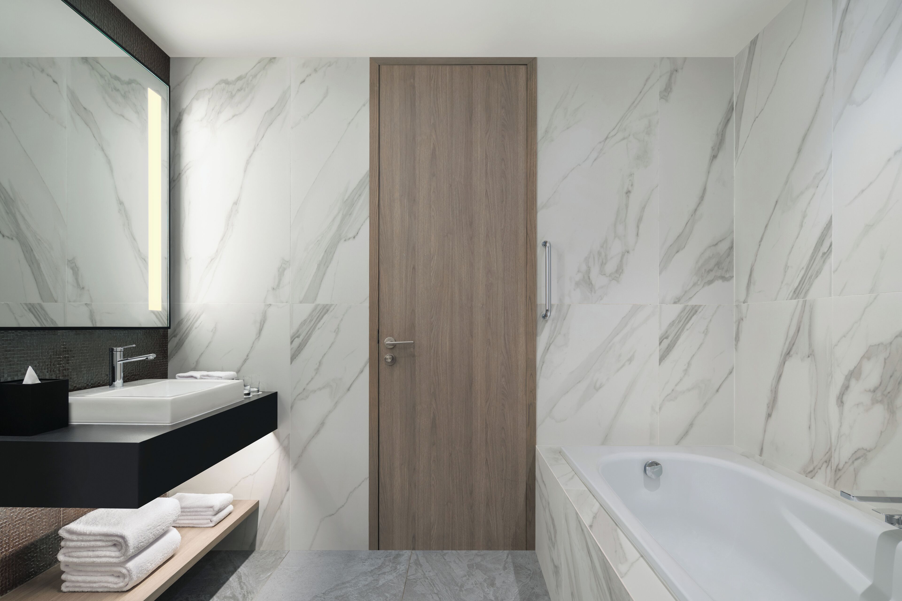 A modern bathroom with a wooden door, a white sink, a bathtub, and marble walls.