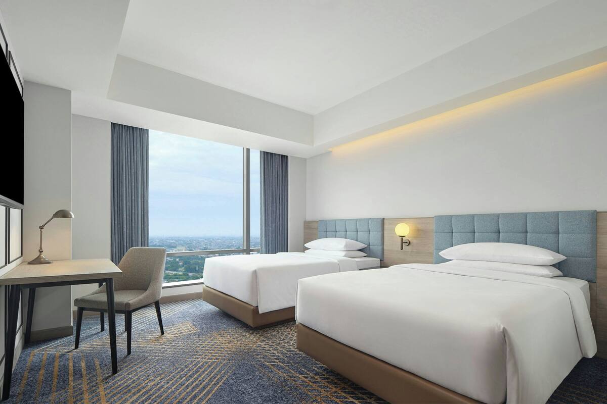 Une chambre d’hôtel avec deux lits, un bureau, une chaise et une grande fenêtre donnant sur la ville.