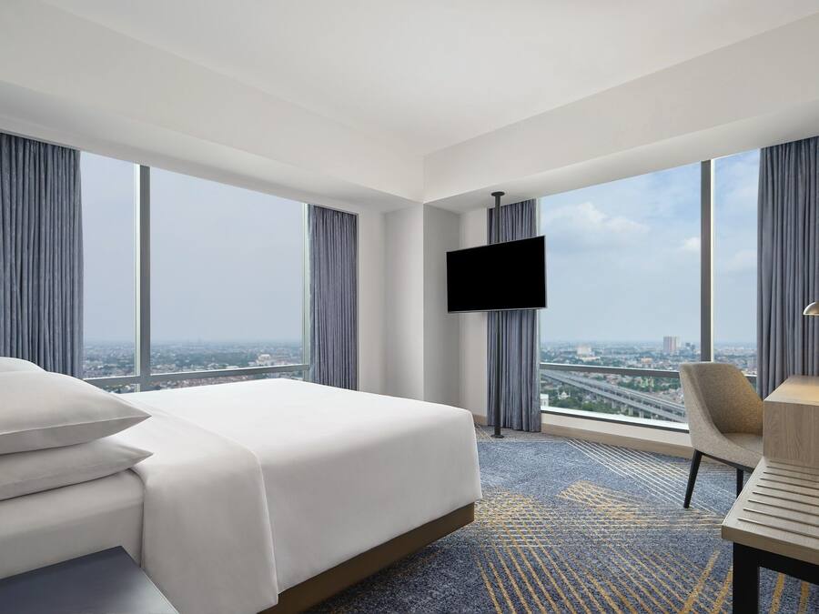 A hotel room with a large bed, a desk with a chair, a flat-screen TV mounted on the wall, and a city view through large windows.