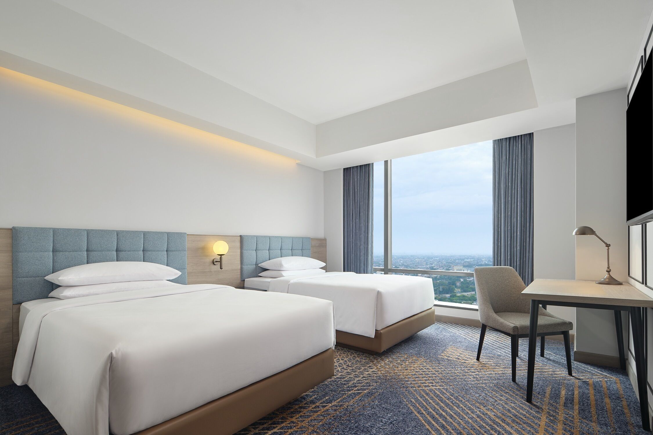 A hotel room with two beds, a desk, a chair, and a large window offering a city view.