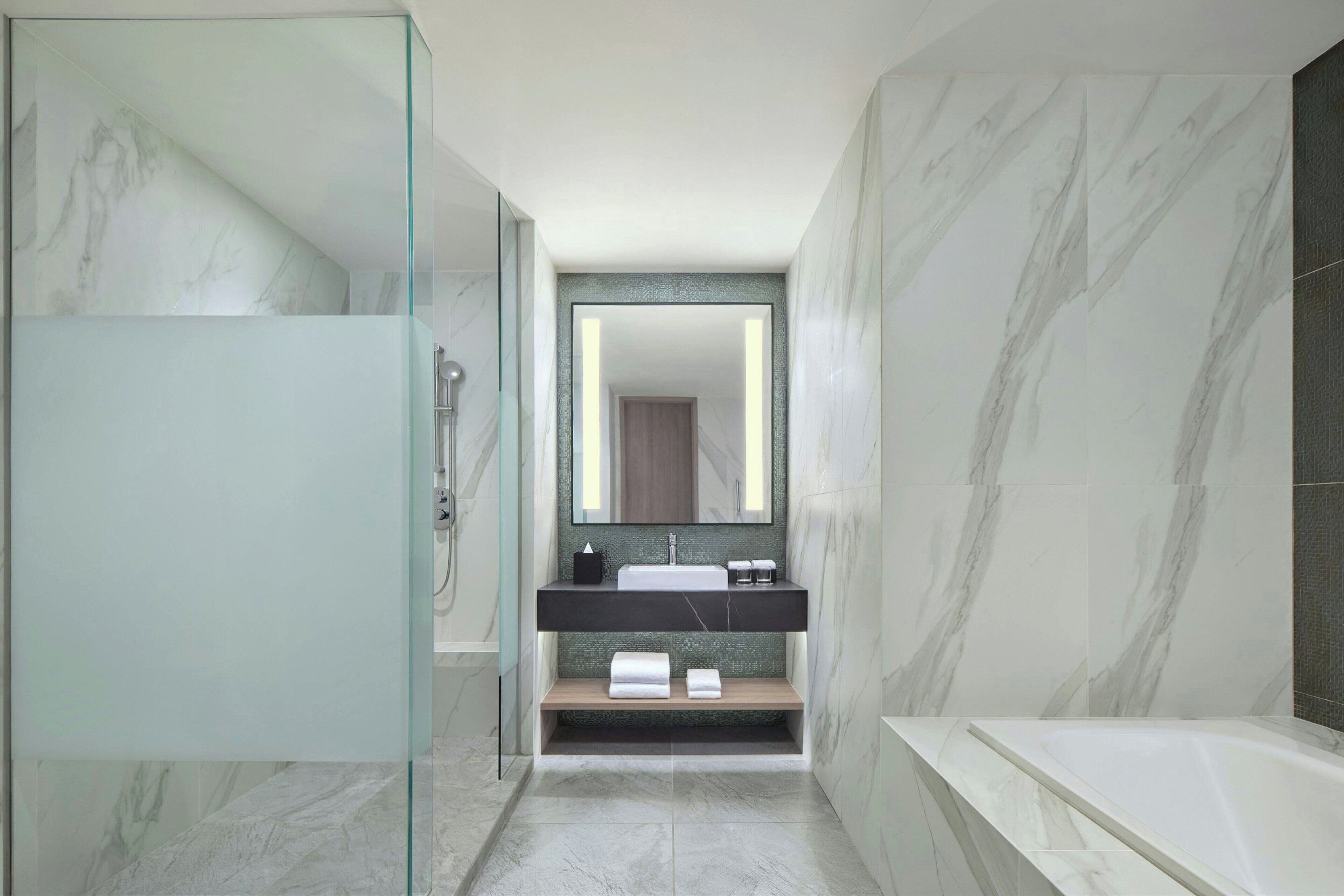 A modern bathroom with a glass shower enclosure, a marble-tiled floor, a built-in sink with a dark countertop, and a freestanding bathtub.